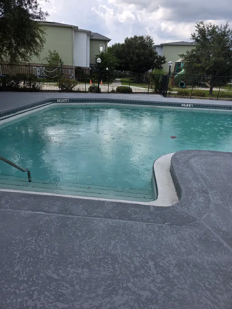 Wide completed view of the gray pool deck, blue water, fence, and community building.