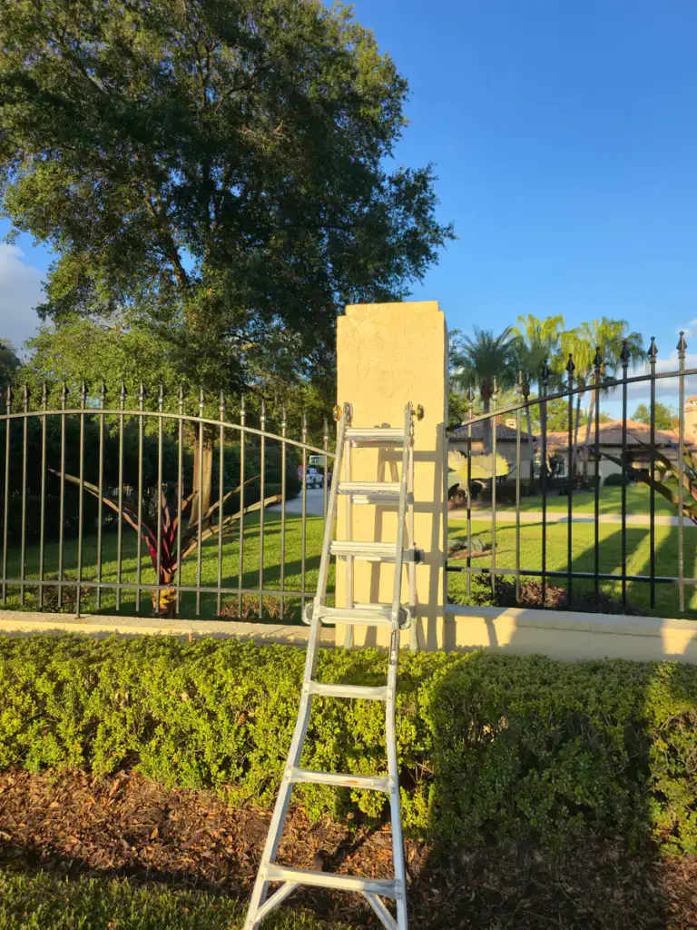 Ladder positioned at a light-colored textured exterior column beside a black metal fence